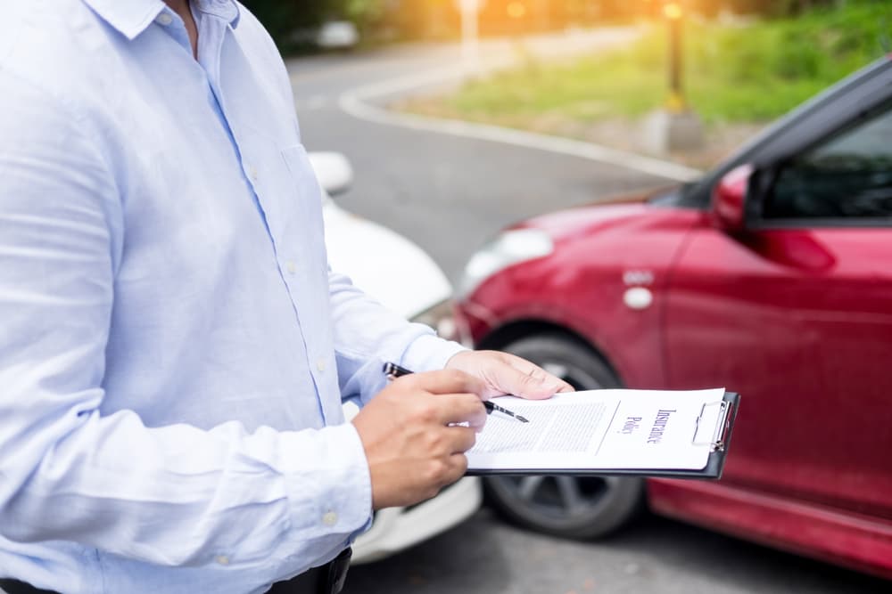 Insurance professional inspecting a damaged car and documenting details on a clipboard as part of the assessment and processing of an accident claim.