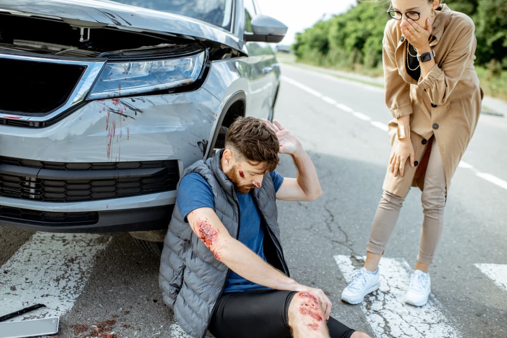 Persona herida en el suelo junto a un coche con una mujer angustiada que se tapa la boca en estado de shock.