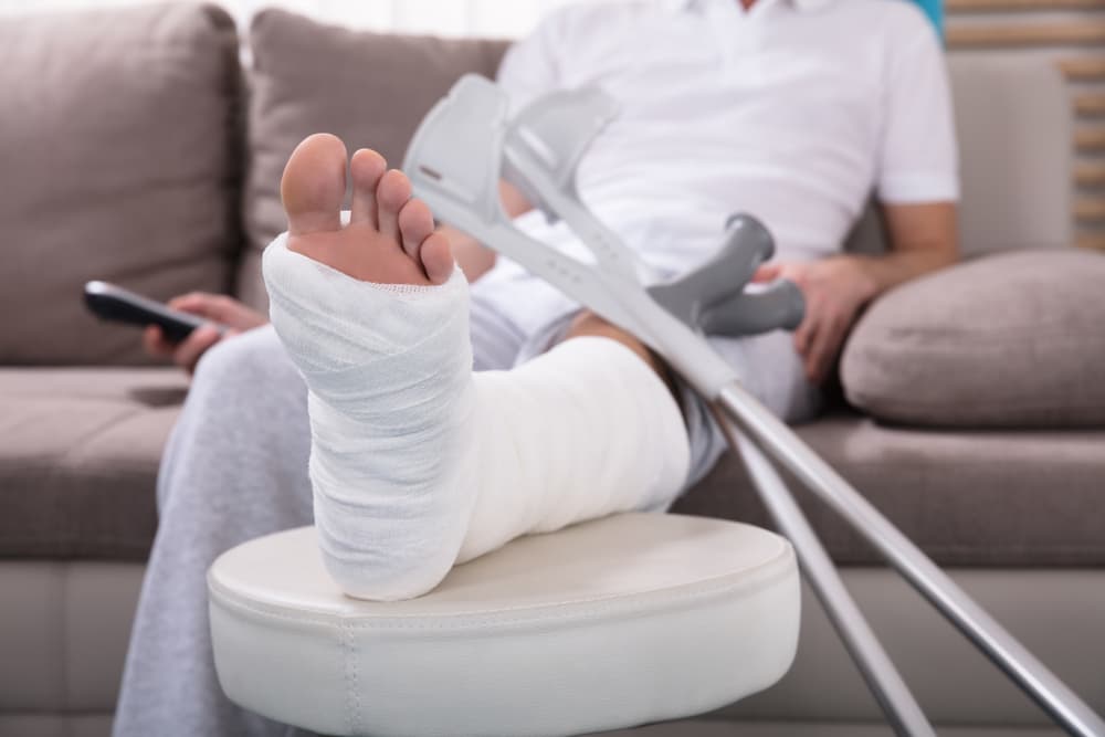 Person with a bandaged leg resting on a footstool with crutches and remote control nearby.