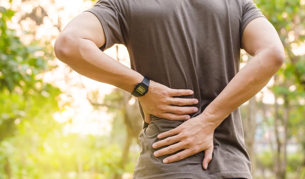 A man outdoors holding his lower back in pain, wearing a smartwatch, with greenery around.