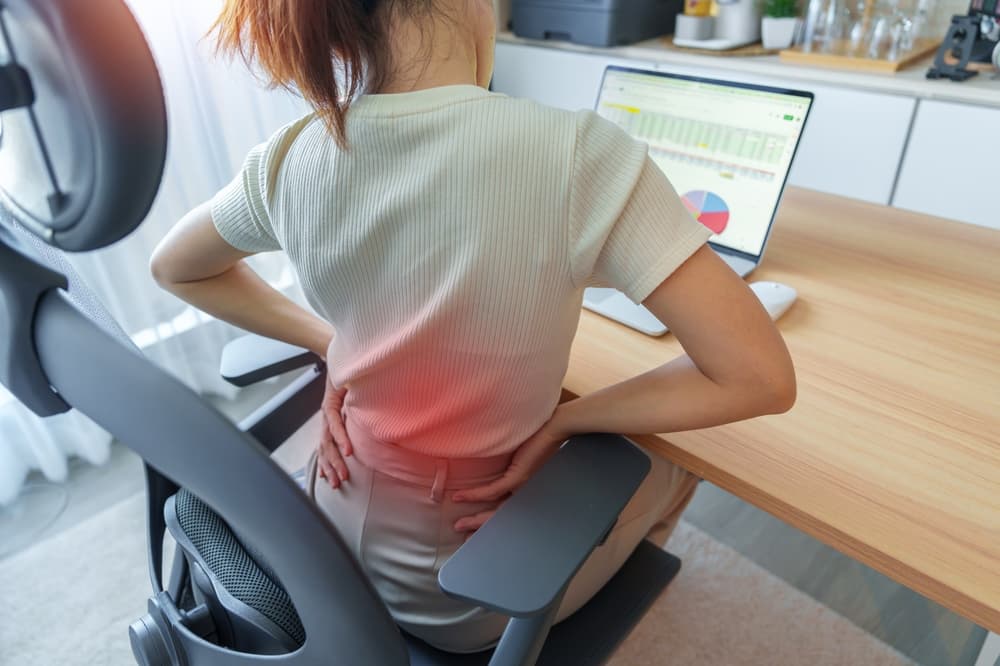 A woman sitting at a desk with a laptop, experiencing back pain and holding her lower back.
