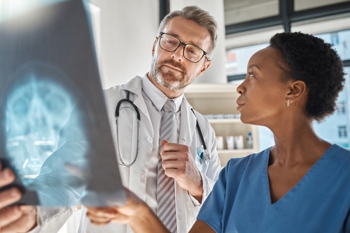 A male doctor and a female nurse in medical scrubs examining an X-ray film together, discussing the patient's condition in a hospital room.